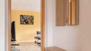 Una habitación moderna con una consola de madera, un espejo grande y una puerta que da a otra habitación con un televisor y un cuadro en la pared.