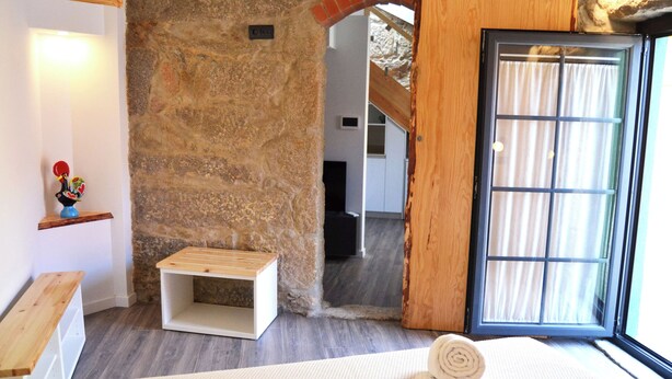 A bedroom with a stone wall, a wooden bench, a bed with a rolled towel, and a window with curtains.