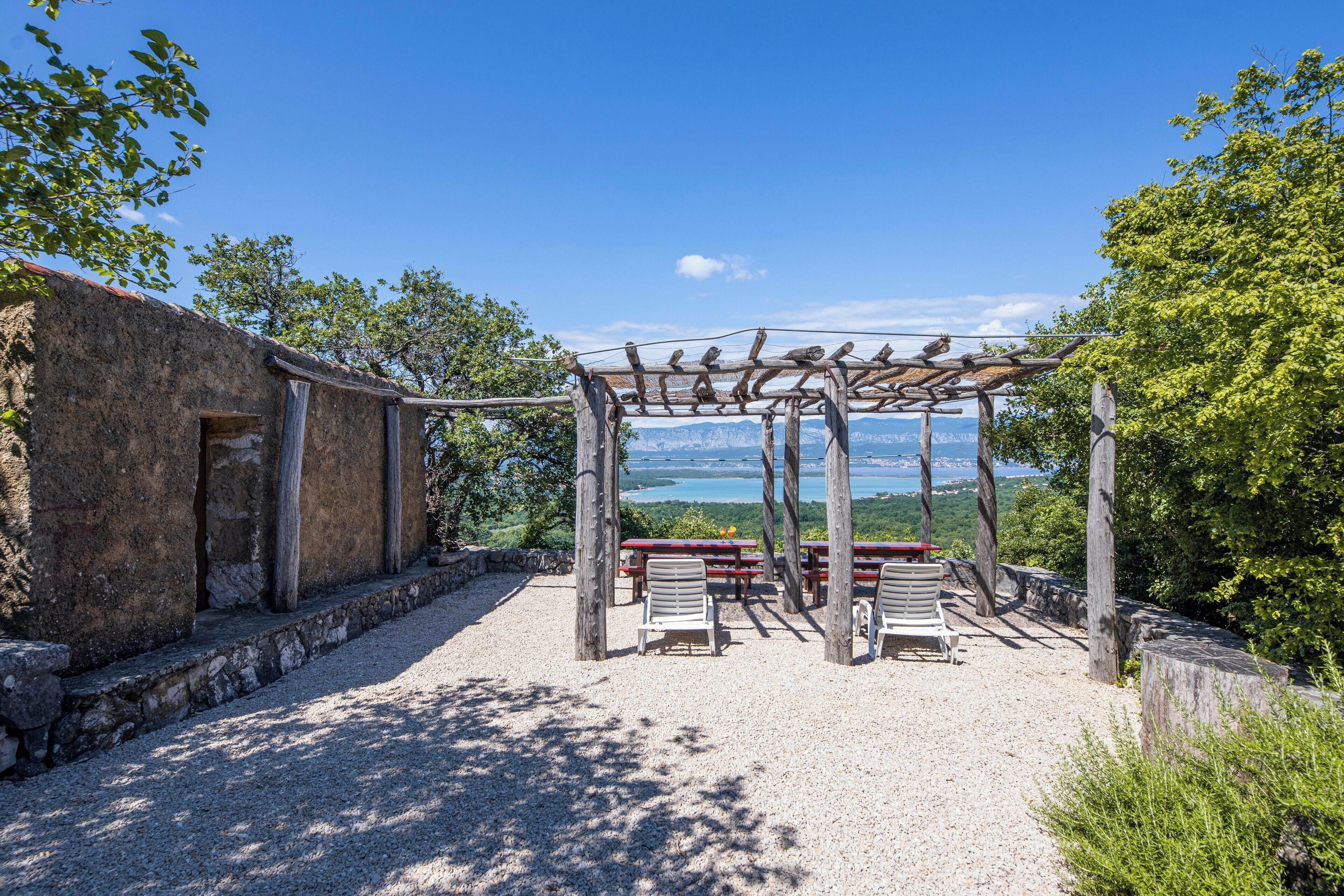 Uma área de estar ao ar livre rústica com pérgola, paredes de pedra e vista para um lago.