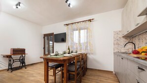 A dining area with a wooden table and chairs, a sewing machine, a television on the wall, and a kitchen with a fruit display. - Lorenzo in Klanice (Haus 2 für 6 Personen) (Dobrinj)