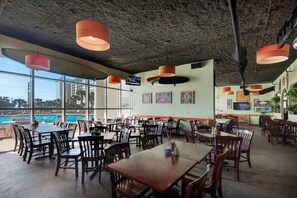 Un vaste espace de restauration intérieur, doté de nombreuses tables et chaises, de grandes fenêtres offrant une vue sur une piscine et des bâtiments, ainsi que d’un plafond avec des poutres apparentes et des luminaires suspendus.