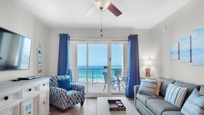 Een woonkamer met een televisie, een bank, een salontafel en een stoel. Er is een balkon met uitzicht op de oceaan.