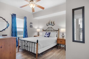 Une chambre à coucher comprenant un lit, un plancher en bois, un ventilateur de plafond, une fenêtre avec des stores, des rideaux, des lampes de chevet et un miroir.