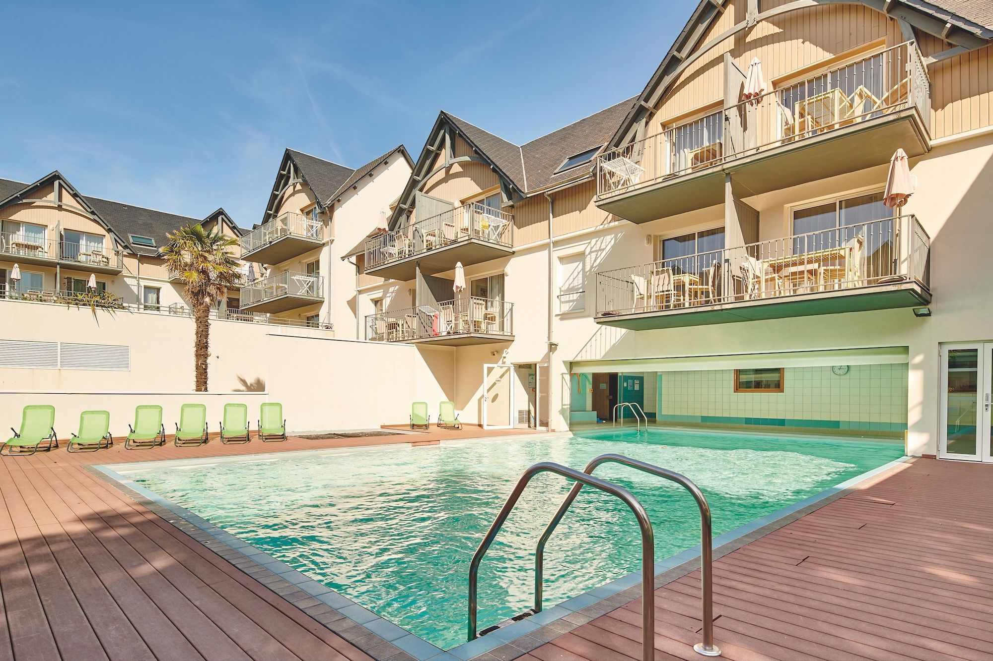 A swimming pool with a wooden deck and green lounge chairs, surrounded by multi-story buildings with balconies.