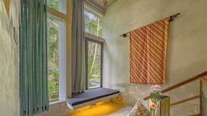 A room with a bench by a window, a patterned wall hanging, and a small table with a vase of flowers. - Gunung An Retreat (Ubud)