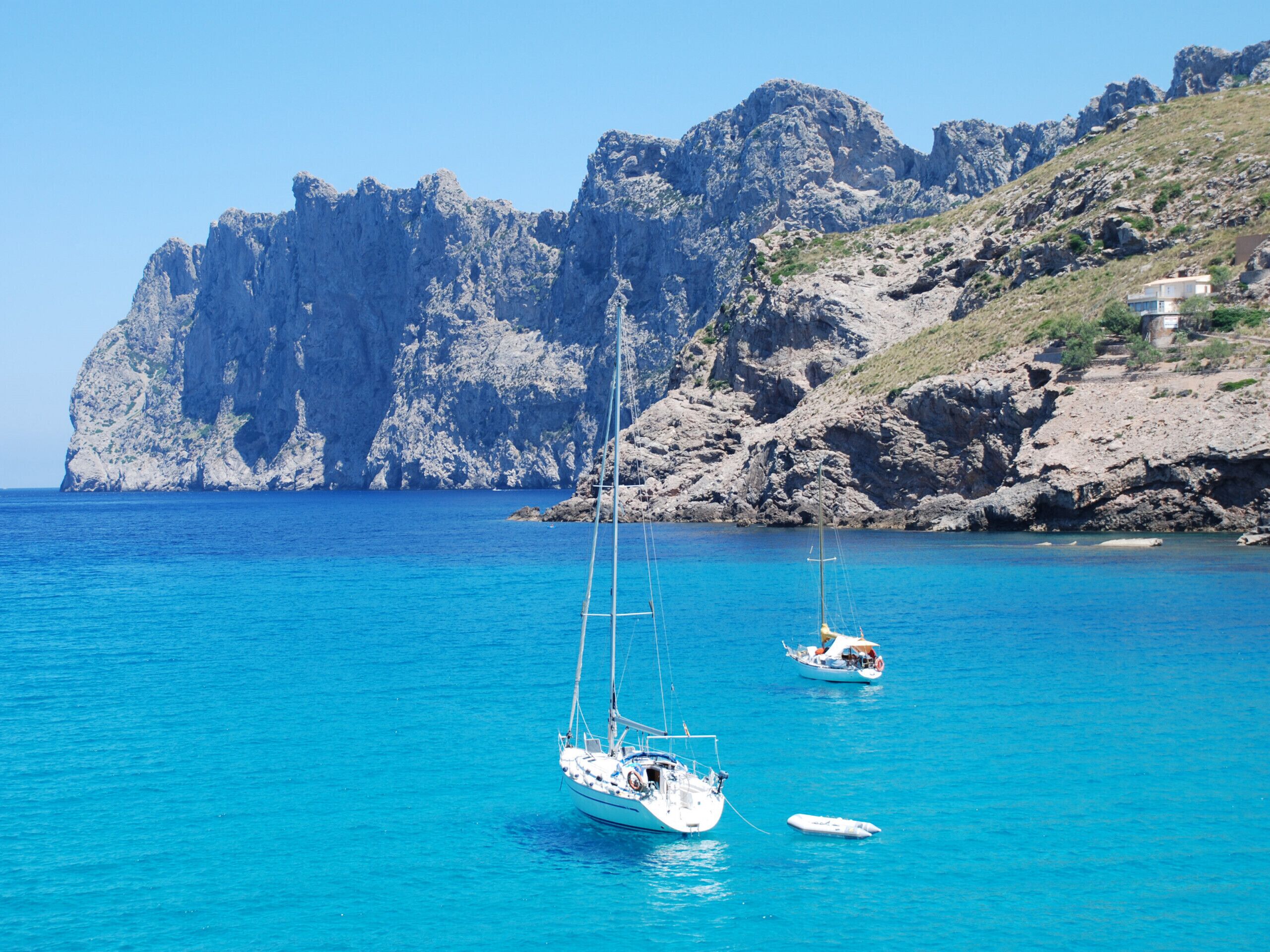 A coastal scene with two sailboats on clear blue water, rocky cliffs in the background, and a building on the hillside.