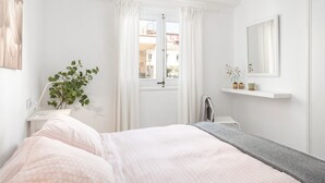 Une chambre à coucher comprenant un lit, une table de chevet, un miroir, une fenêtre avec des rideaux et une vue sur les bâtiments à l’extérieur.