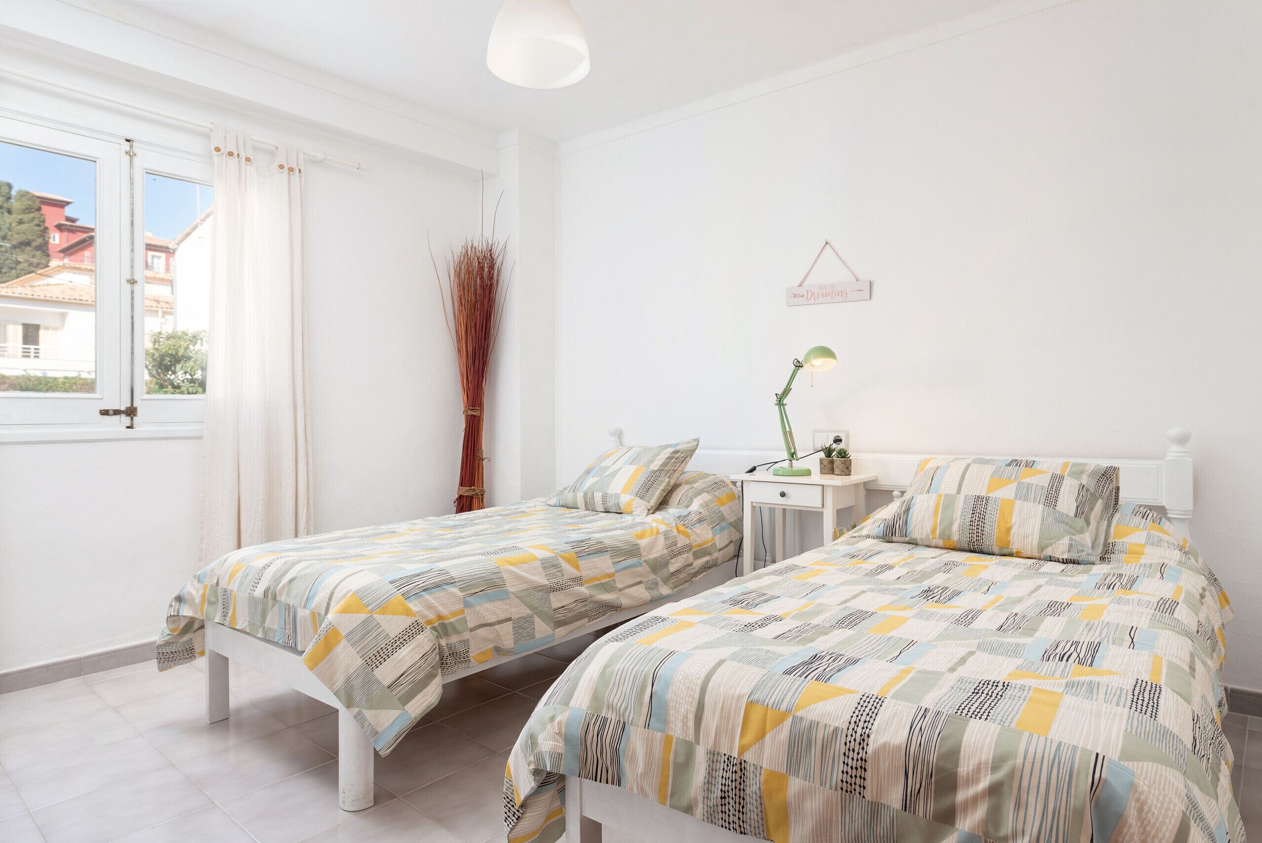 A bedroom with two beds, a bedside table with a lamp, and a window with curtains.