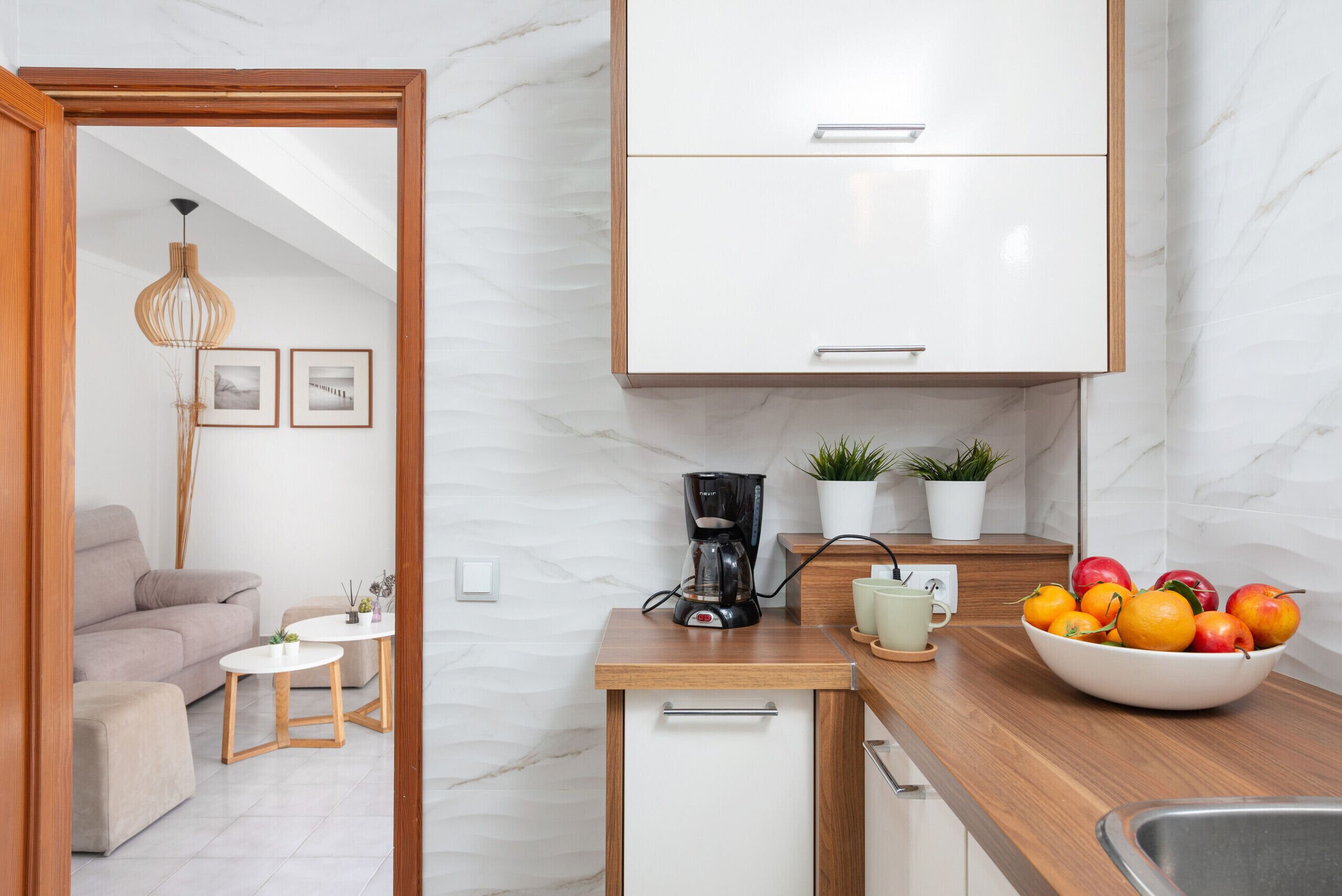 Una cocina moderna con armarios blancos, una cafetera, un frutero y un pequeño comedor con un sofá y mesa.