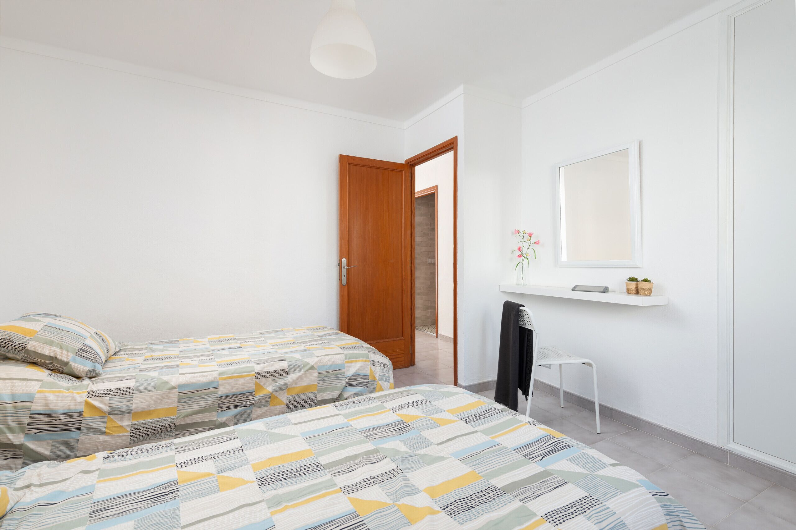 A bedroom with two beds, a wooden door, a white chair, a small table with a mirror, and a shelf with a plant.