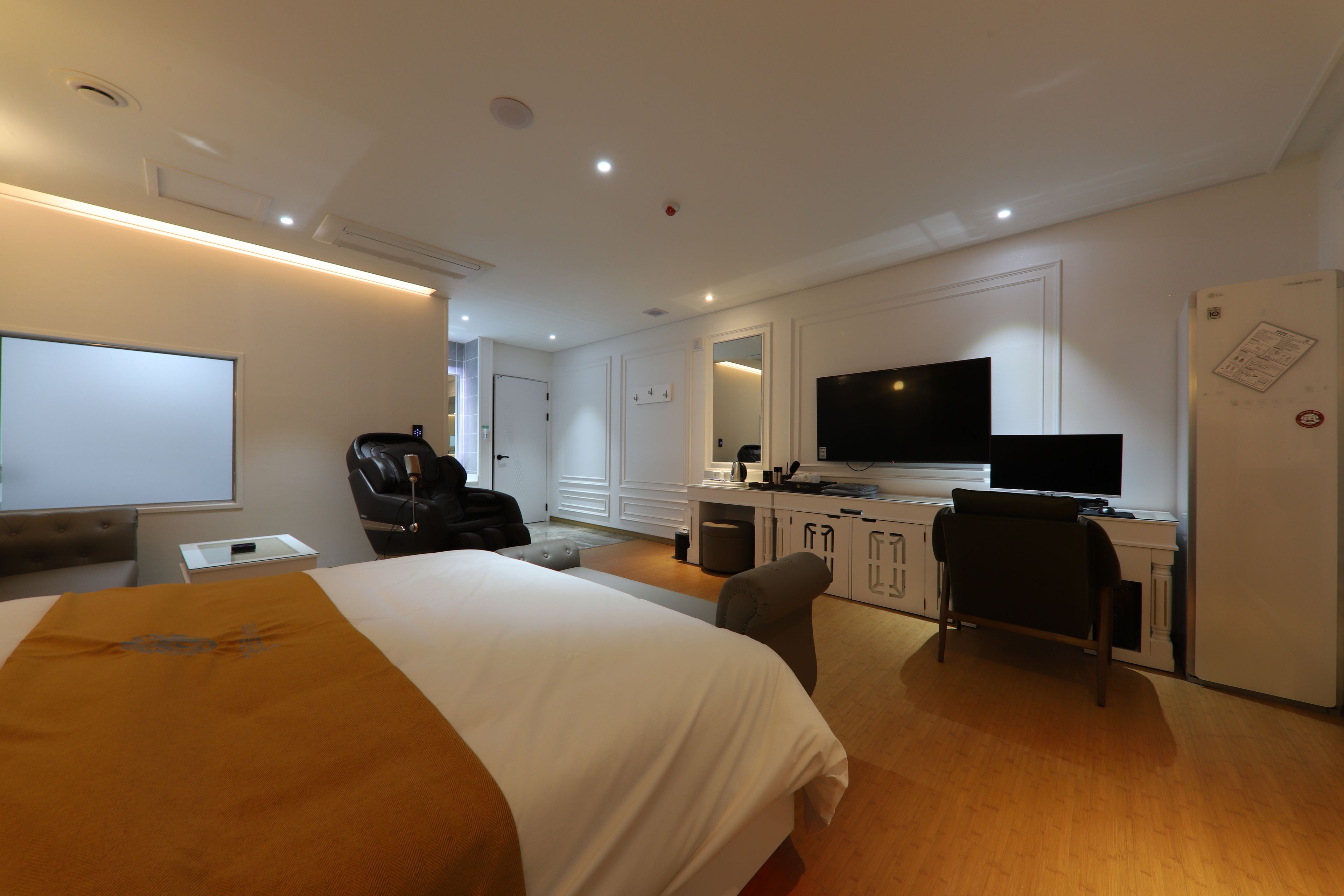 A hotel room with a bed, a desk with a flat-screen TV, a chair, a desk lamp, a whiteboard, and a refrigerator.