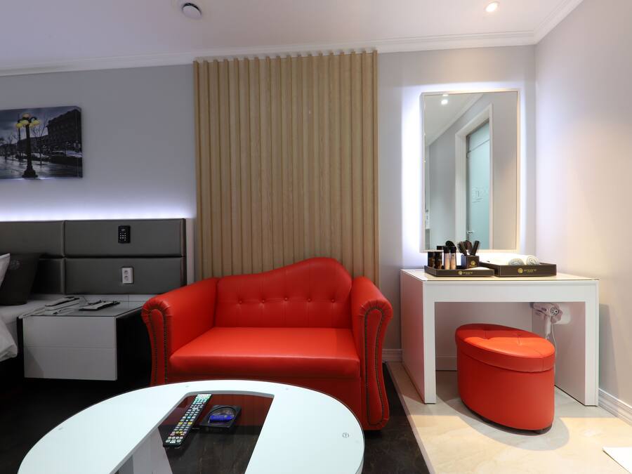 A modern hotel room with a red sofa, a white desk with a mirror, a small red ottoman, and a bed with a grey headboard.