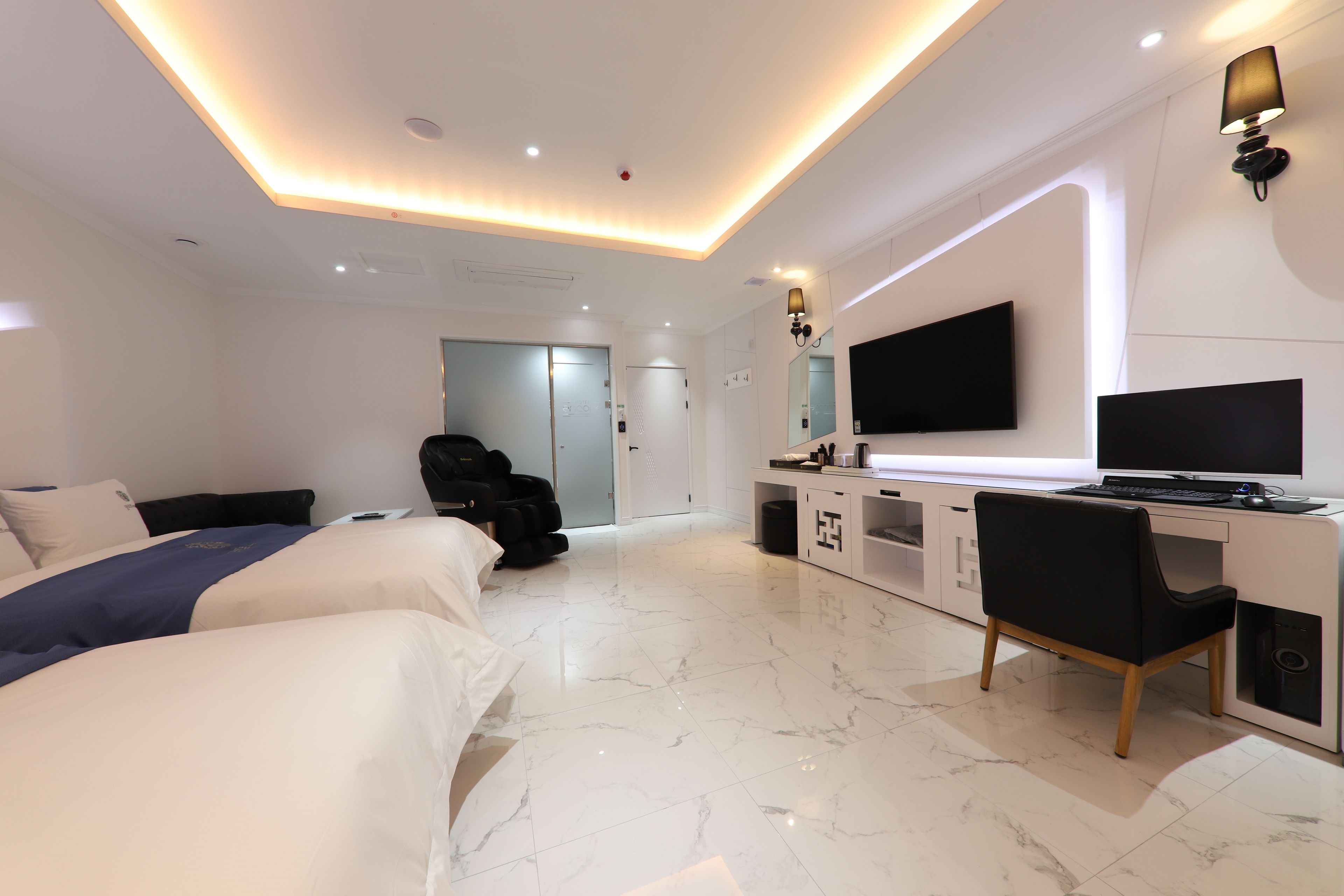 A modern hotel room with a large bed, a desk with a computer, a TV, and a massage chair.