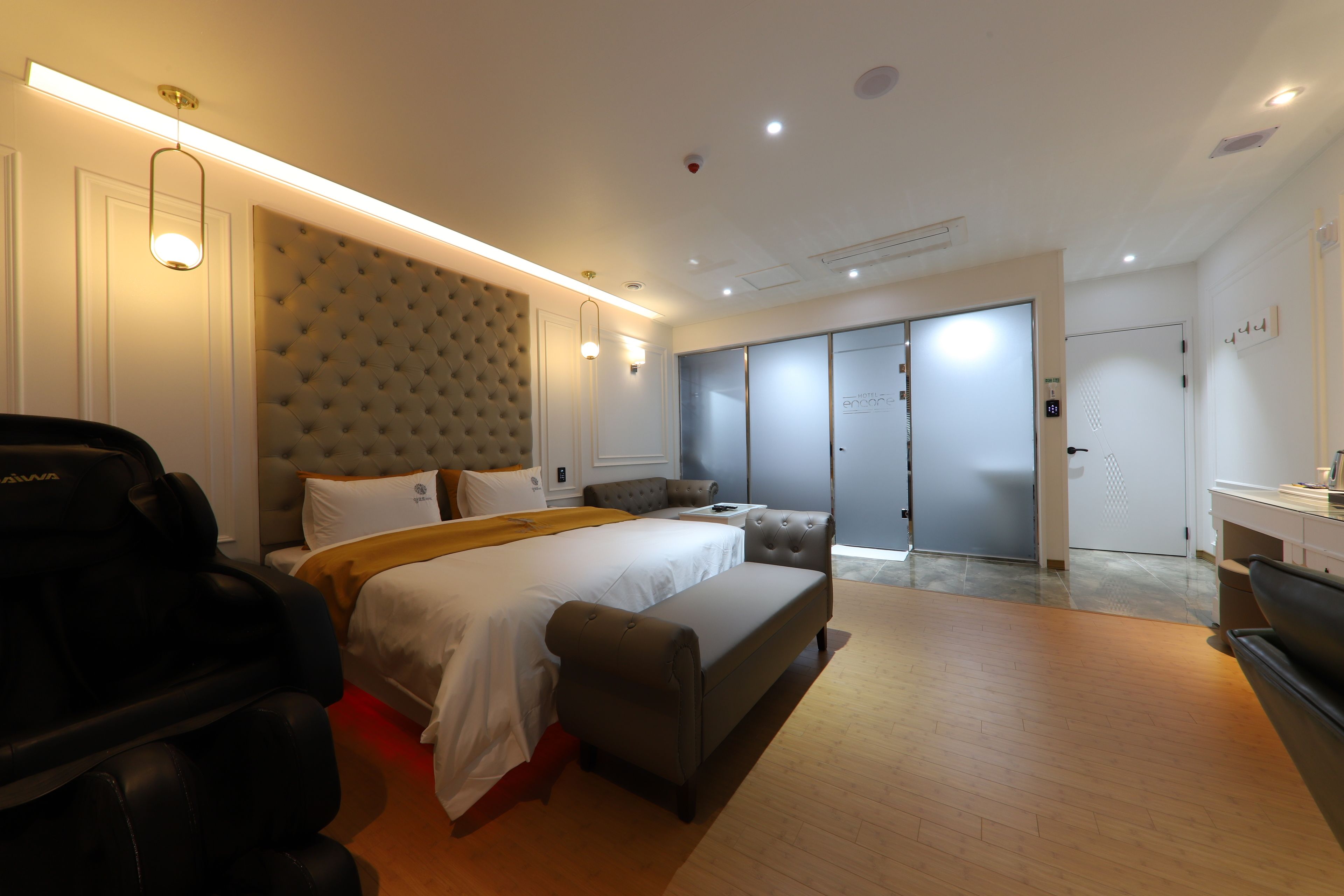 A modern hotel room with a large bed, a massage chair, a grey sofa, and a sliding door closet.