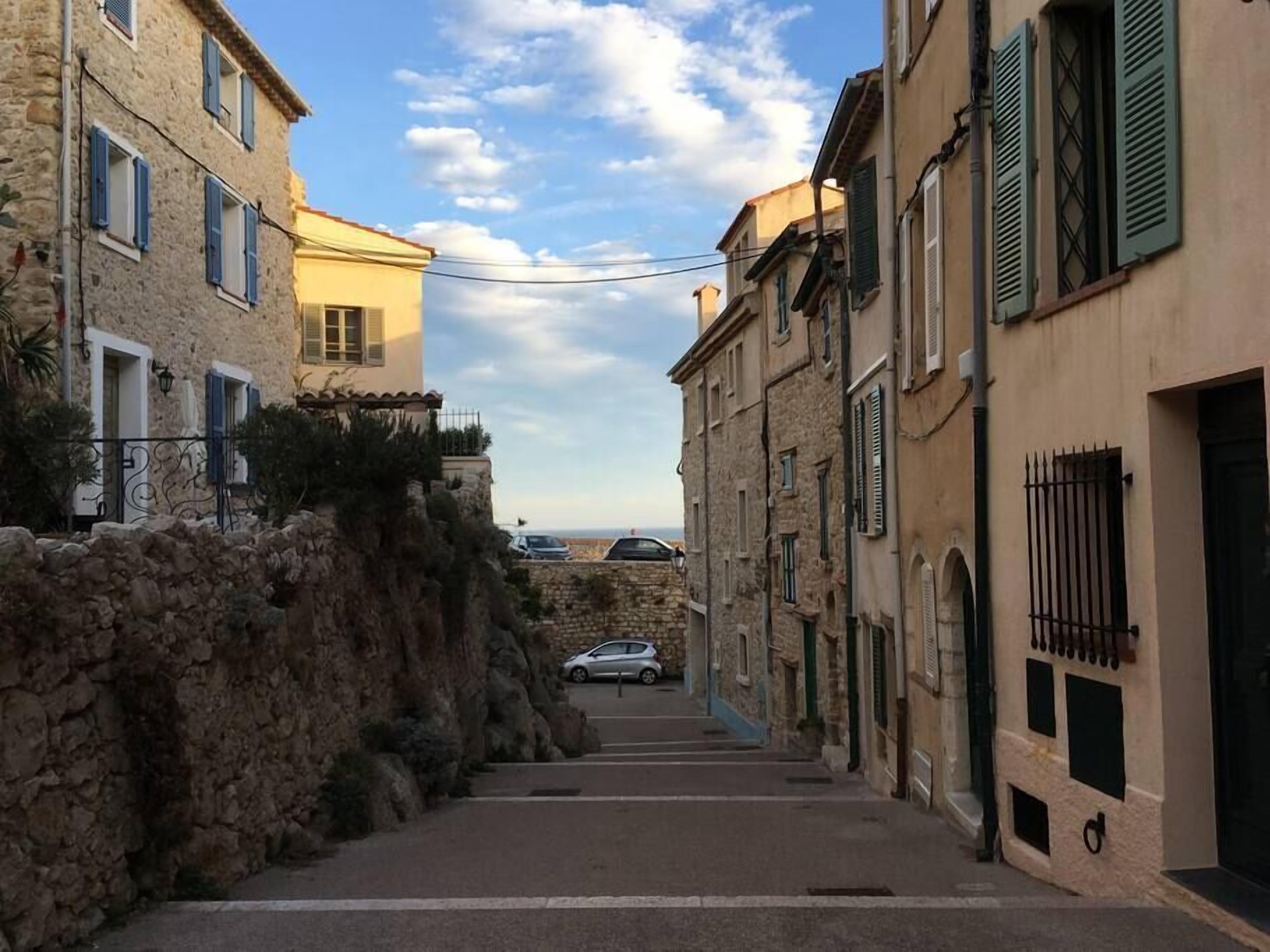 A narrow street flanked by stone buildings with shutters, leading to a distant view.