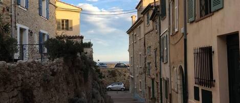 A narrow street flanked by stone buildings with shutters, leading to a distant view.