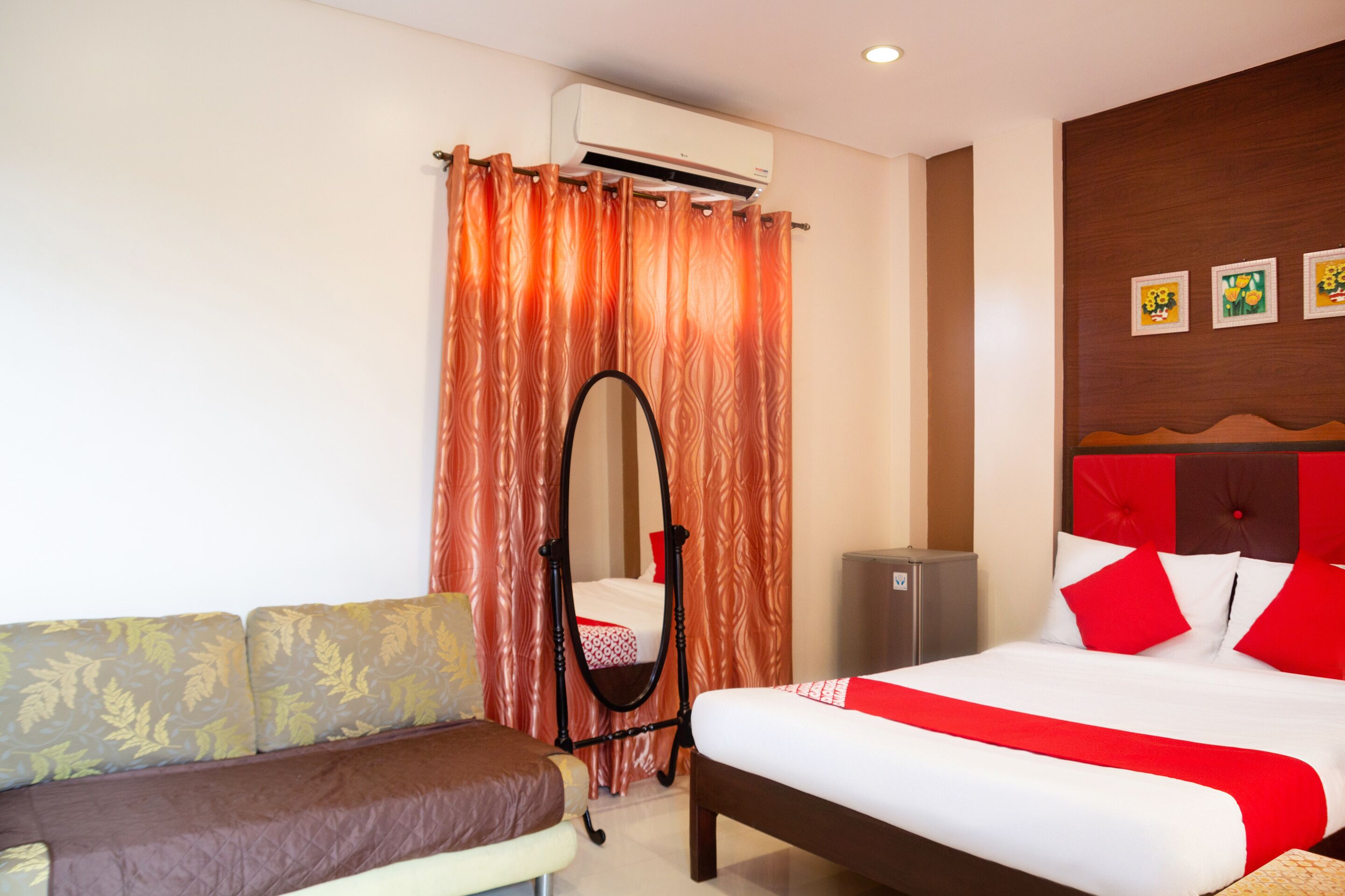 A hotel room with a bed, a sofa, an air conditioning unit, a mirror, and a wall with framed pictures.