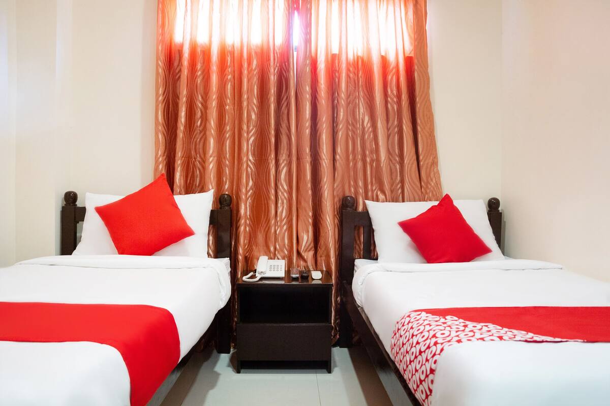 A hotel room with two beds, each with white bedding and red pillows. There is a nightstand between the beds with a phone and a small table.