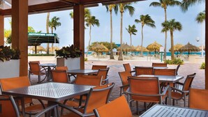 An outdoor seating area with tables and chairs, overlooking a beach with palm trees and a sign reading "ACTIVITIES AREA".