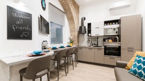 Une cuisine moderne avec une arche en briques, un tableau noir sur lequel est écrit le message « Bienvenue à tous », et un coin repas avec des tabourets de bar.