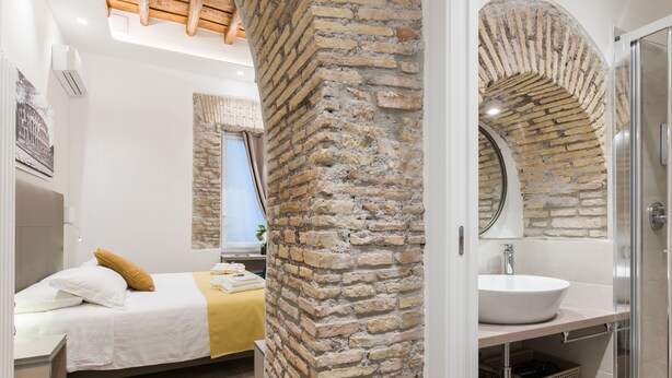 Une chambre moderne avec un mur en briques, un lit avec une literie blanche et un plaid jaune, une table de chevet et une fenêtre avec des rideaux.