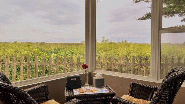 Un patio cubierto con mobiliario de mimbre, una pequeña mesa con un florero y una caja, y vistas a un campo cubierto de hierba a través de una valla de madera.