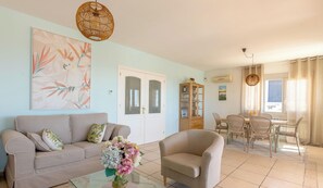 Un salon comprenant un canapé, un fauteuil et une table basse en verre ornée de fleurs. On y trouve également un coin repas avec une table et des chaises, un buffet et un tableau accroché au mur.