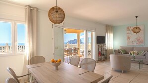 Ein Zimmer mit Esstisch, Stühlen, einer Couch, einem Fernseher und einem Balkon mit Meerblick.