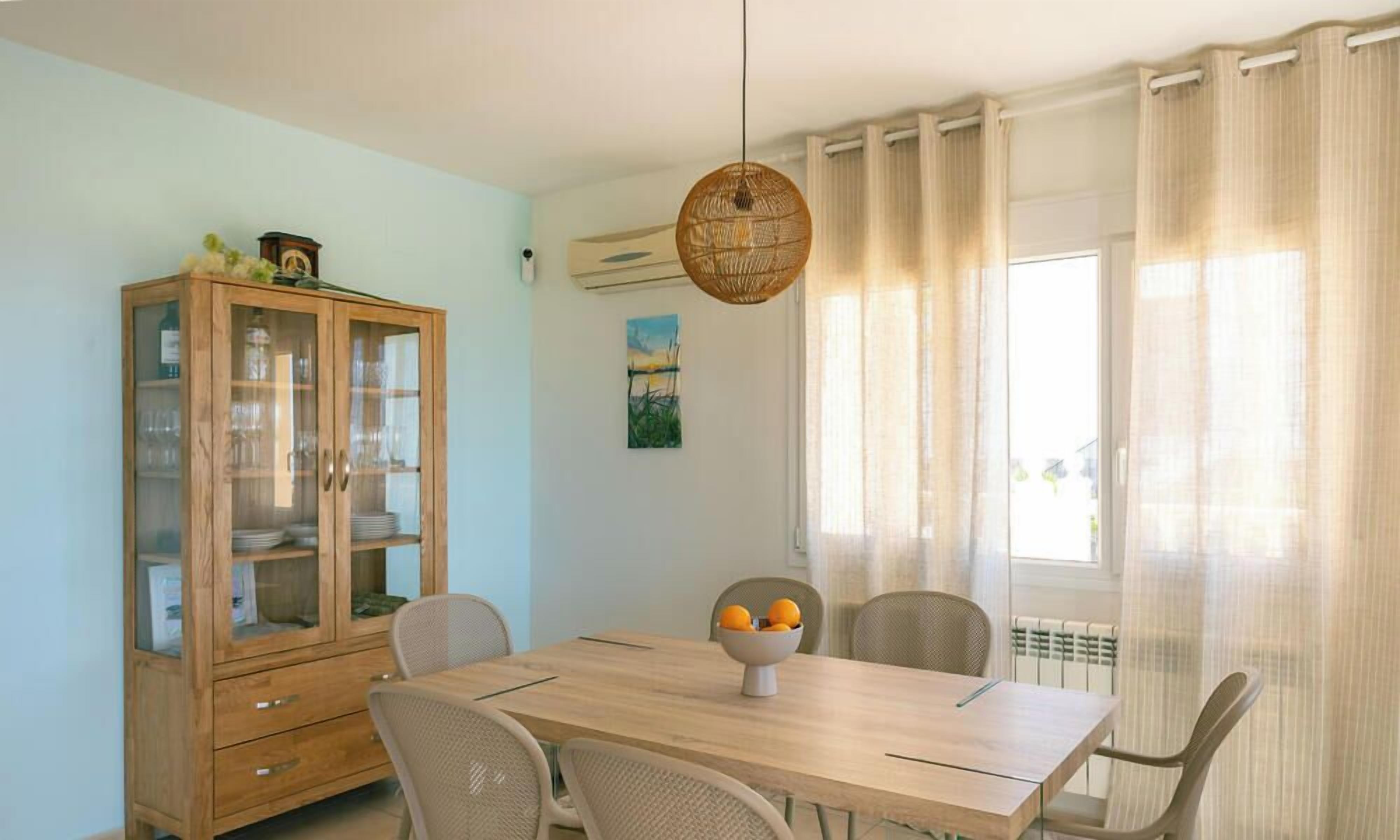 A dining area with a wooden table, chairs, a cabinet with glass doors, a hanging light fixture, and a window with curtains.
