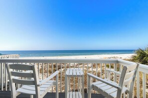 A beach view from a wooden deck with white chairs and a small table.