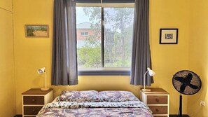 A bedroom with a bed, two bedside tables, a standing fan, a window with curtains, and a wall-mounted picture. - Riverwood - nestled beside the Wooli River (Wooli)