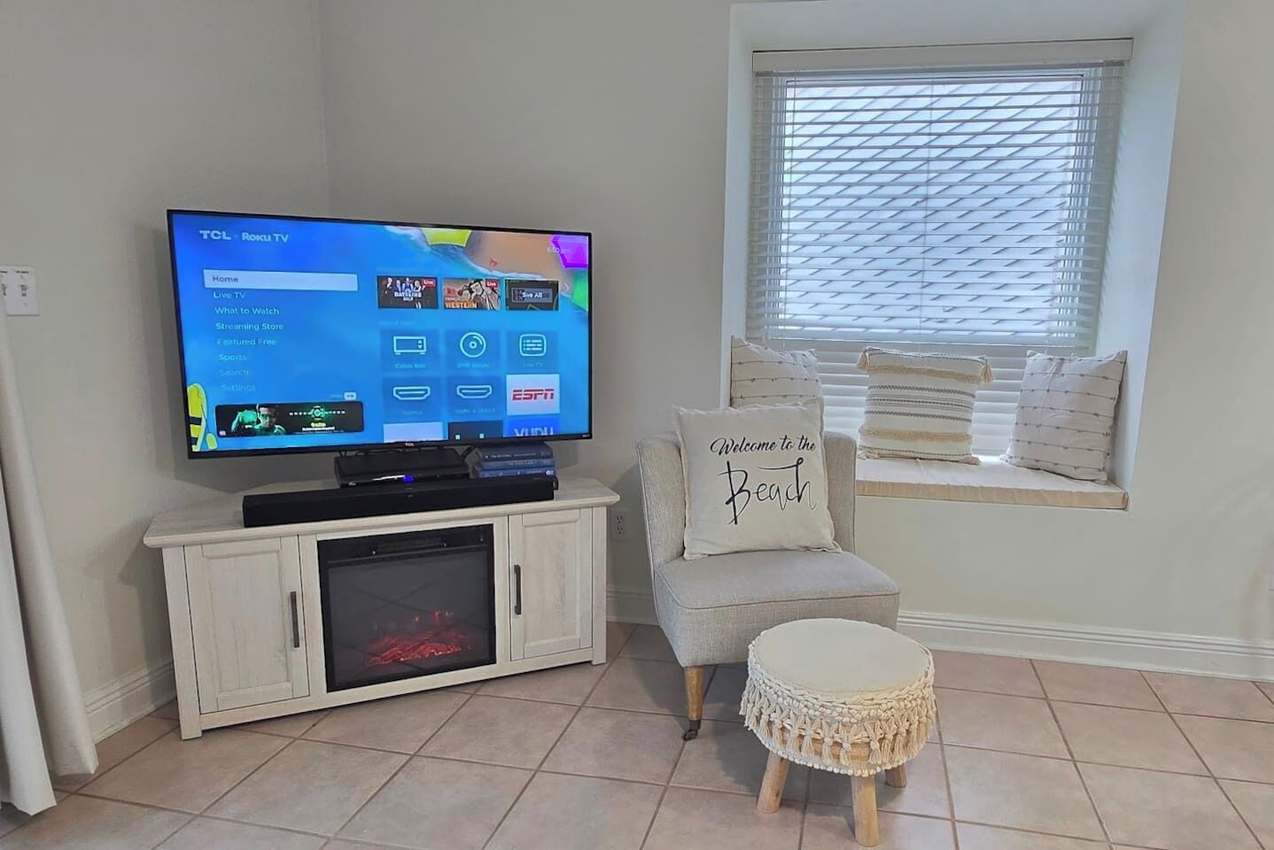 Una sala de estar con televisor, un mueble bajo, un banco junto a la ventana con cojines y un taburete decorativo.