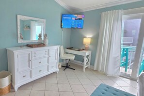 Une pièce avec une commode blanche, un bureau avec une lampe, un téléviseur fixé au mur et une vue sur l’extérieur par une fenêtre.