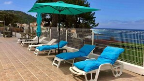 A seaside terrace with white lounge chairs and blue cushions, a turquoise umbrella, and a view of the ocean. - 4-person apartment in the heart of the pevani estate (CALCATOGGIO)