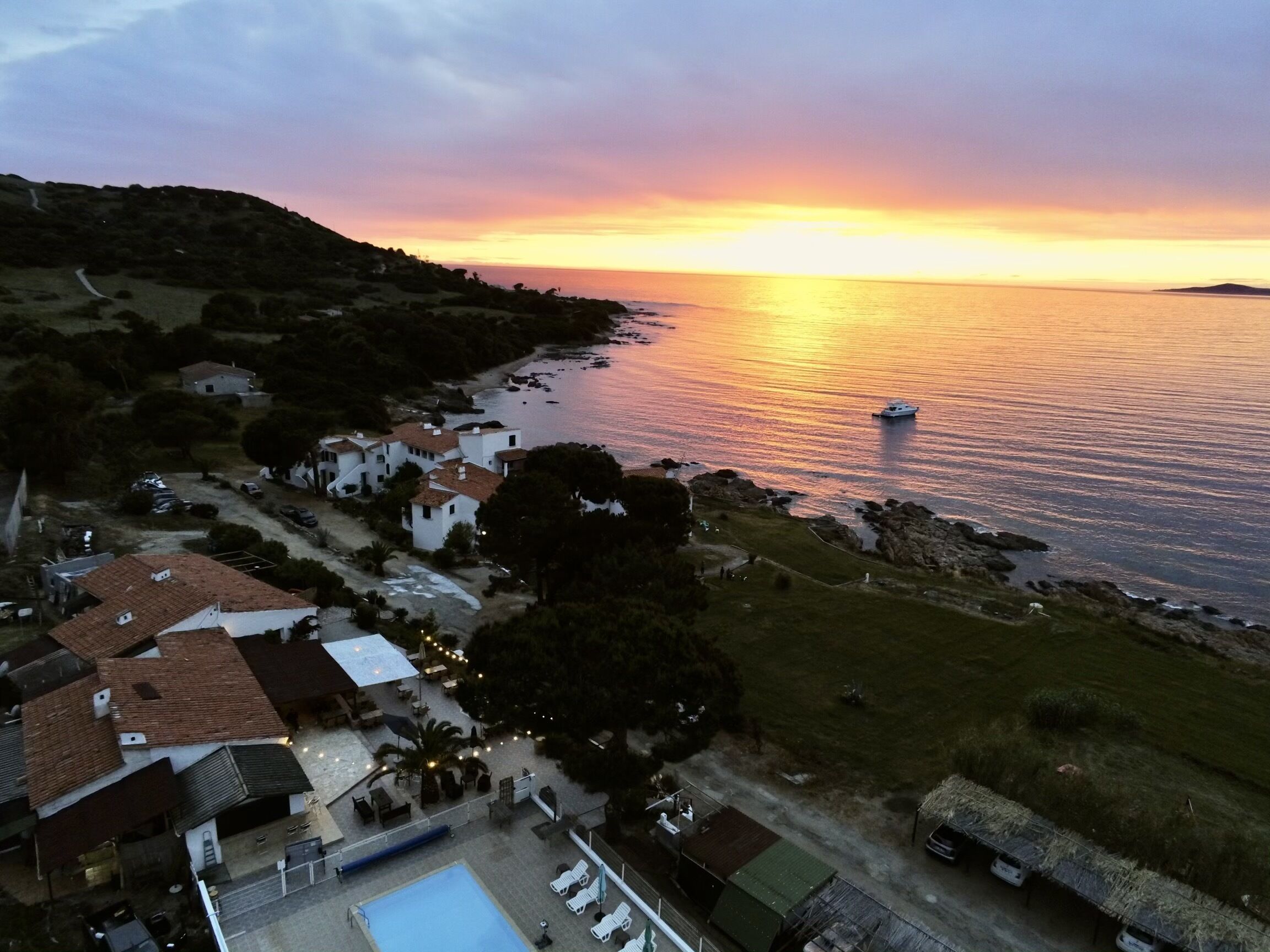 A coastal property with a swimming pool, a small boat docked near the water, and a view of the sunset over the sea.
