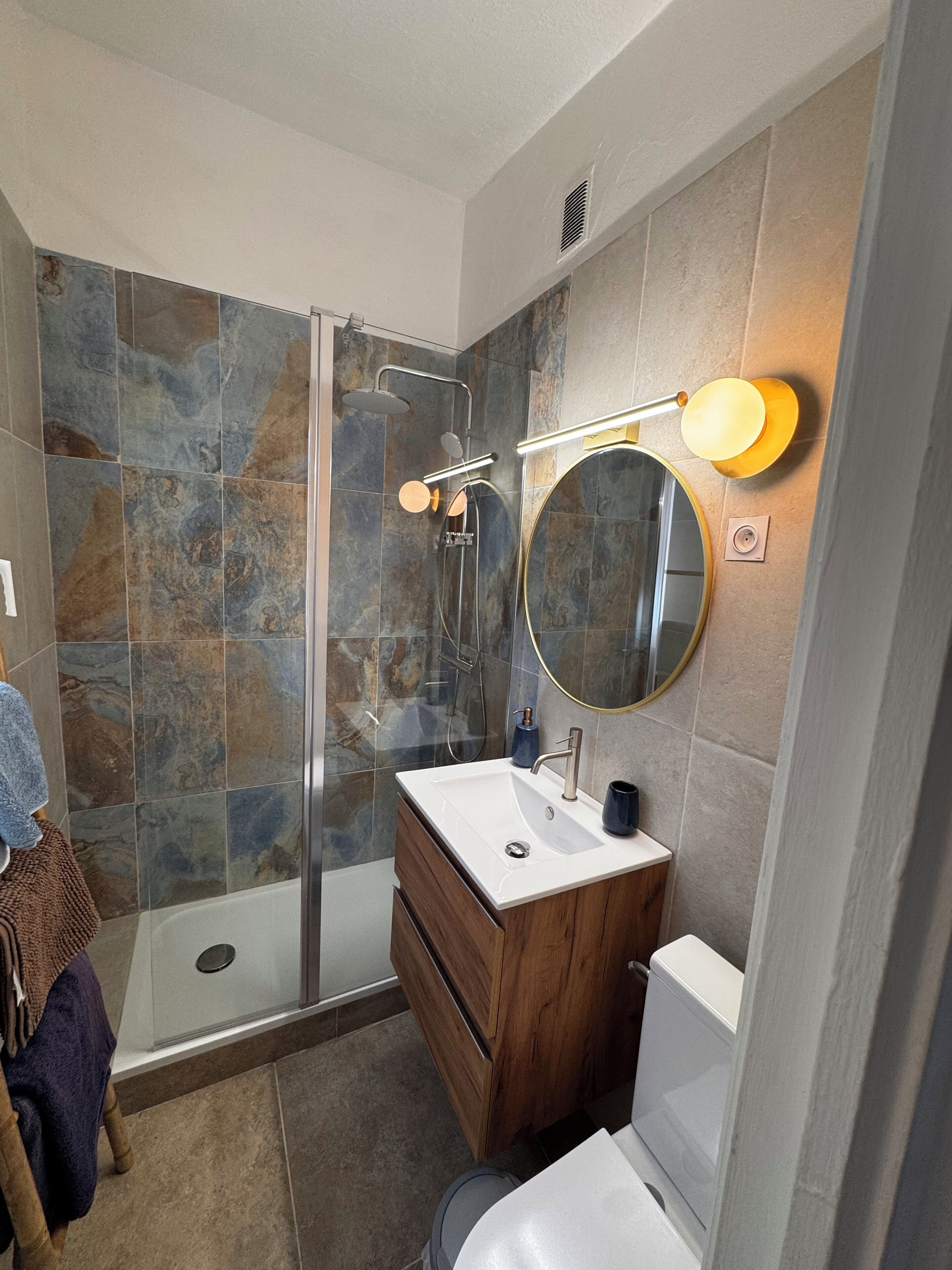 Een moderne badkamer met een glazen douchecabine, een houten wastafelmeubel en een ronde spiegel met verlichting.