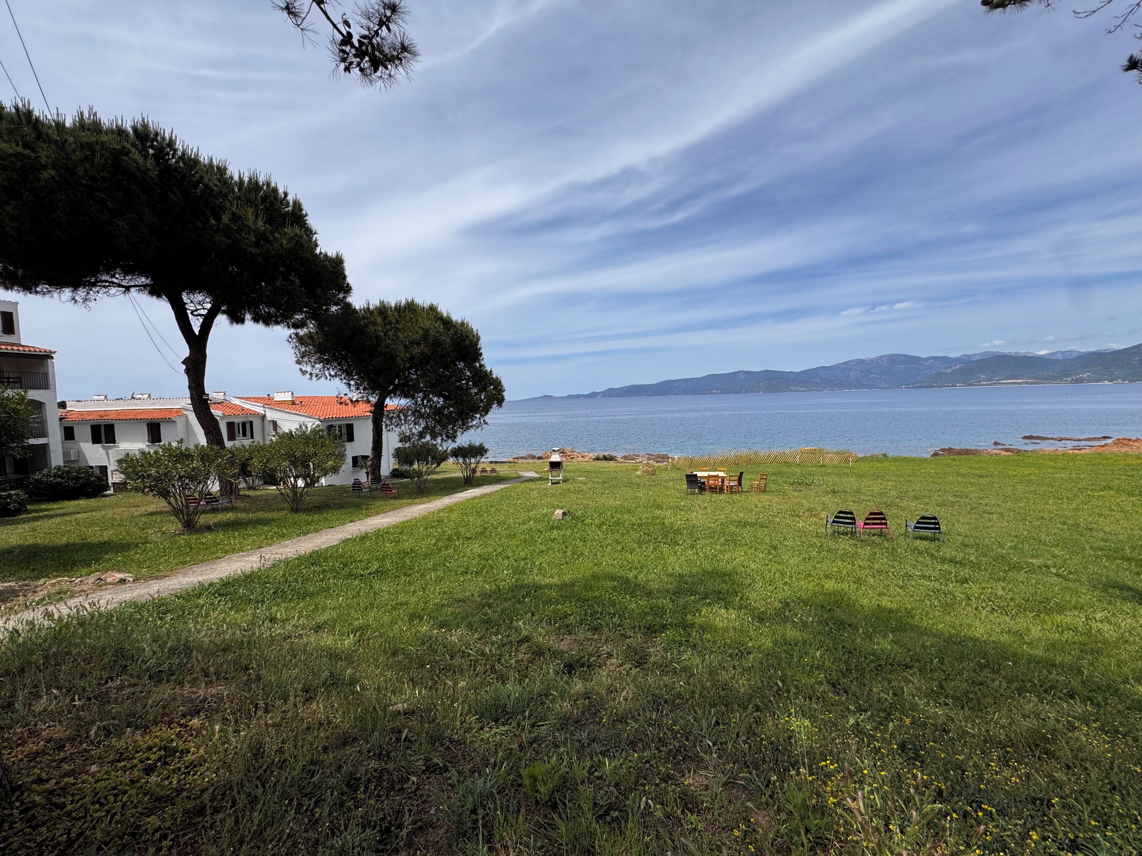 Une zone herbeuse avec un sentier menant à un bâtiment doté d’un toit recouvert de tuiles rouges, un grand arbre et une vue sur la mer.
