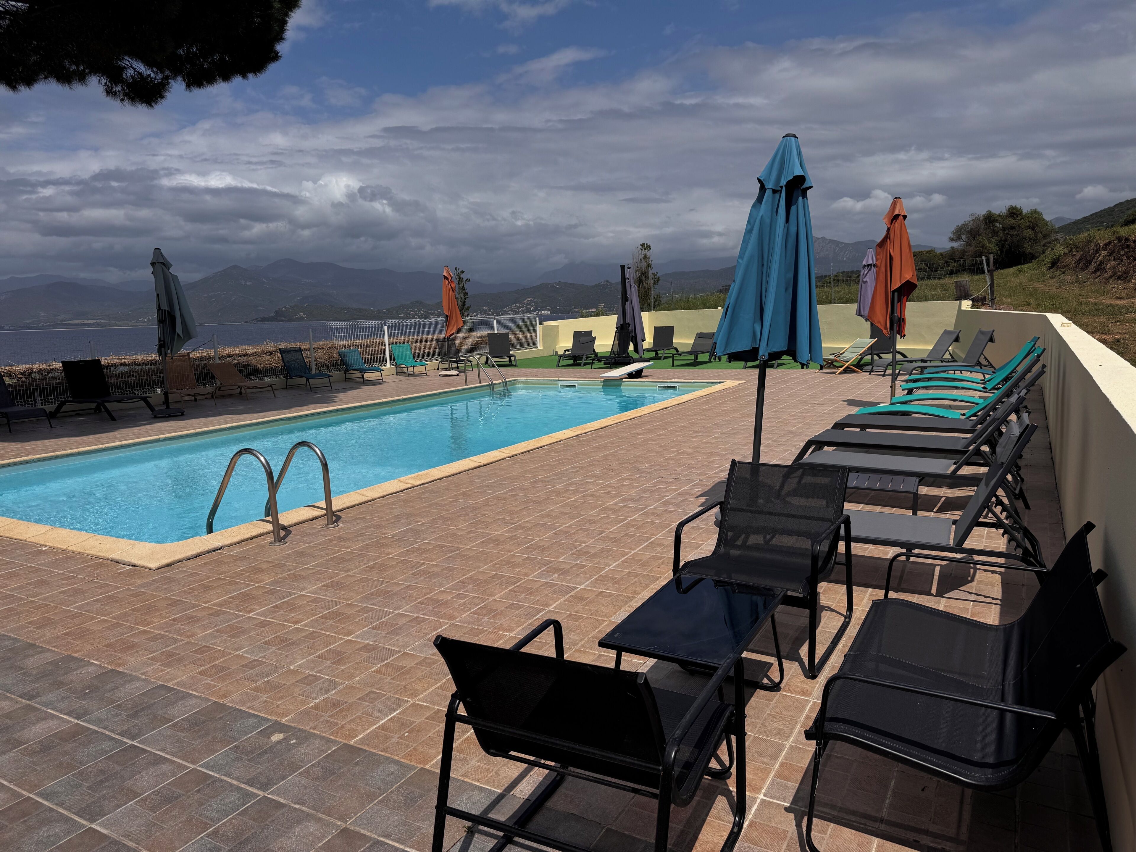Una piscina con lettini e ombrelloni, circondata da una recinzione e montagne sullo sfondo.