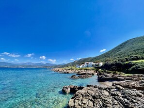 A coastal landscape with clear blue waters, rocky shores, and a white building with multiple balconies overlooking the sea.
