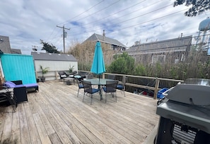 A wooden deck with a table and chairs, a blue umbrella, and a grill. - Fire Island Beach Haven (Ocean Beach)