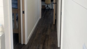 Ein schmaler Korridor mit holzverkleideten Wänden und dunklem Holzboden. Eine Tür links führt in einen Raum mit einem Etagenbett. Ein Spiegel befindet sich über einem Schreibtisch mit Stuhl an der Wand.