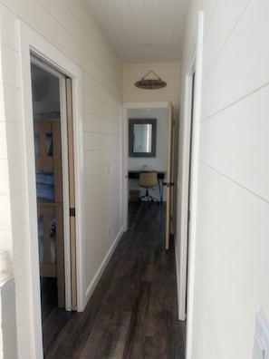 A narrow hallway with wooden panel walls and a dark wood floor. A door on the left leads to a room with a bunk bed. A mirror hangs on the wall above a desk with a chair. - Fire Island Beach Haven (Ocean Beach)