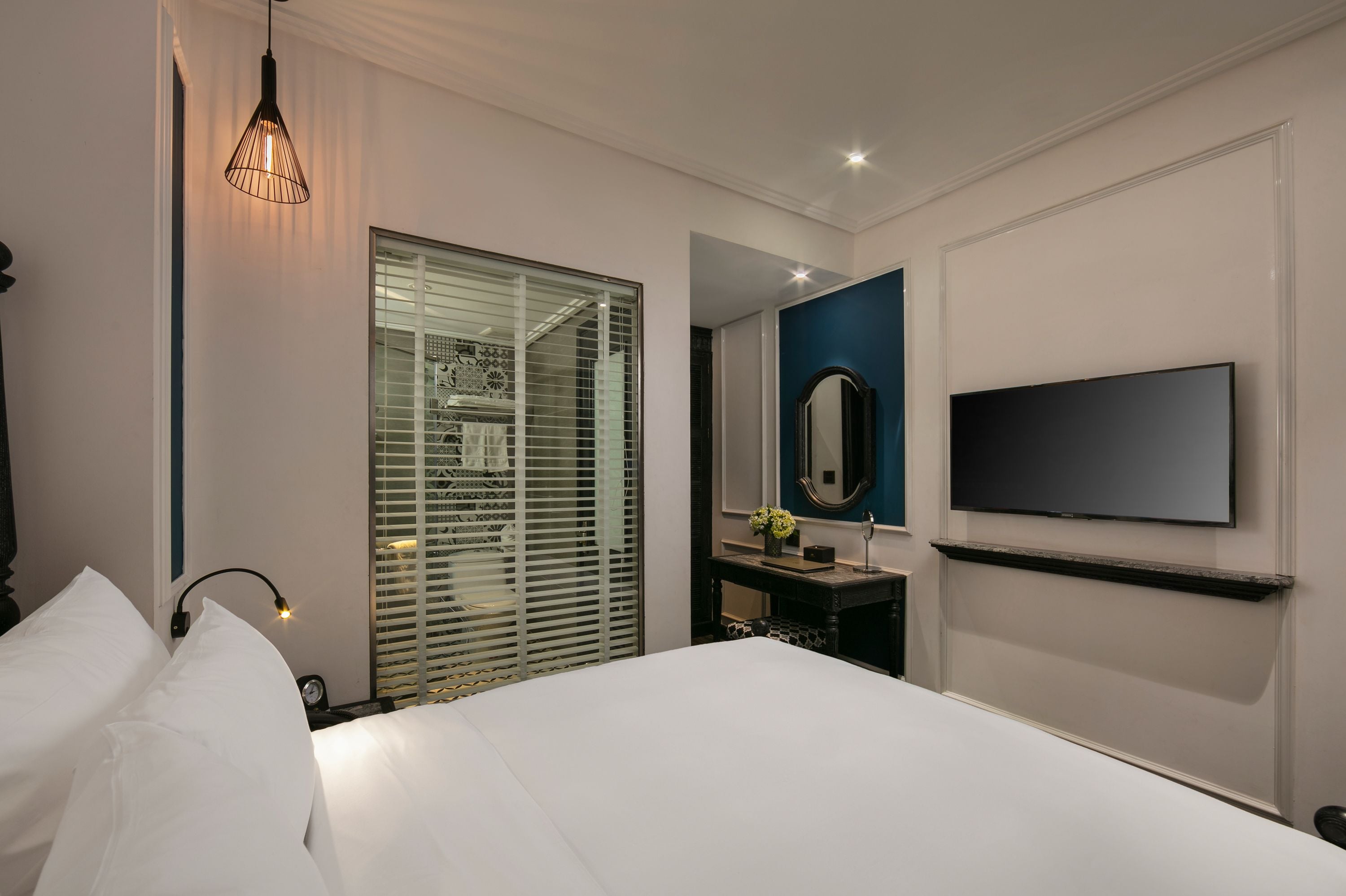 Una habitació d'hotel moderna amb un llit gran, una televisió penjada a la paret, una taula de nit i una finestra amb persianes.