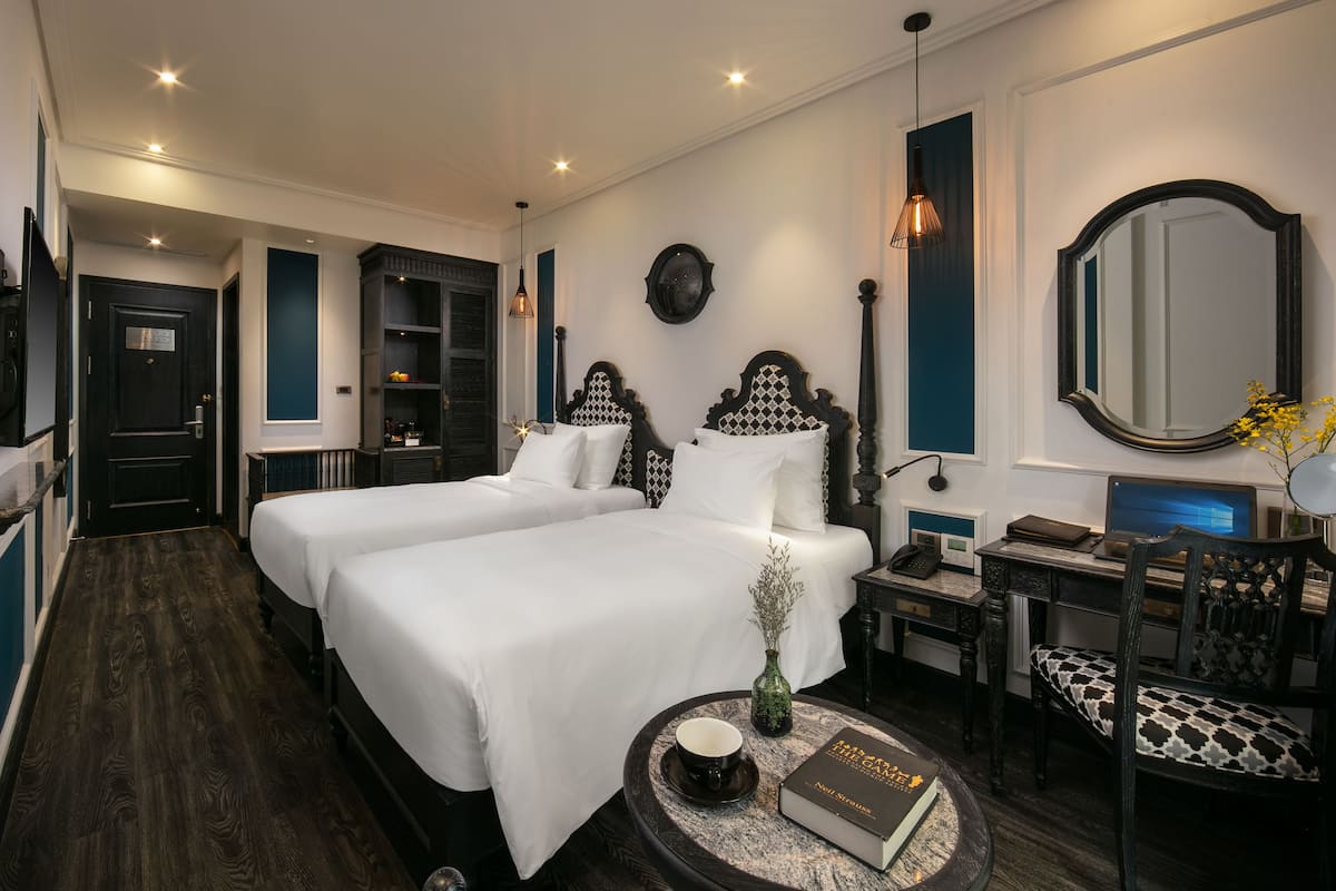 A hotel room with two beds, a desk, a chair, a round table with a book and a cup, and a mirror on the wall.