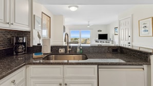 A modern kitchen with granite countertops, white cabinetry, a stainless steel sink, and a view of the ocean through large windows.