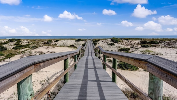 A wooden boardwalk leading to a beach with sandy dunes and sparse vegetation.