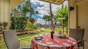 Une table dressée pour deux personnes, recouverte d’une nappe à motifs floraux rouges et blancs, donnant sur un jardin tropical avec des palmiers et un ciel dégagé.