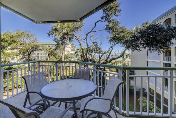 A balcony with a round table and chairs, overlooking a landscaped area with trees and a building in the background. - 2316 Windsor II - Beautiful 2 Bedroom Villa with beautiful views! (Hilton Head Island)
