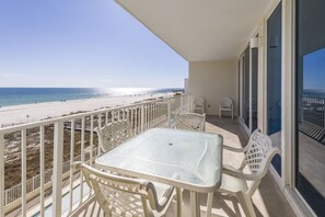 A balcony with a glass table and white chairs, overlooking a beach with a clear sky.