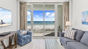 Une pièce dotée d’une grande porte-fenêtre coulissante donnant sur un balcon avec vue sur la plage. On y trouve un fauteuil bleu à motifs, un canapé gris et une petite table avec une lampe.